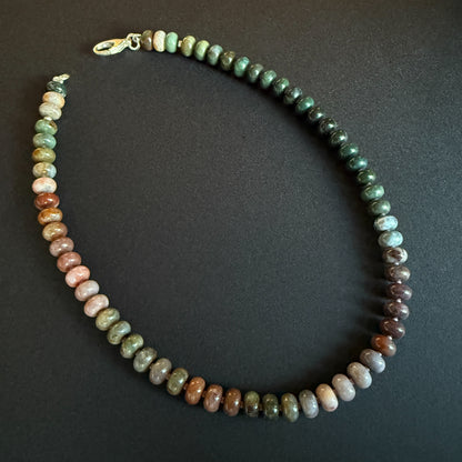 A close-up of a gemstone necklace with smooth, rounded beads in a variety of natural hues, secured with a metallic clasp.