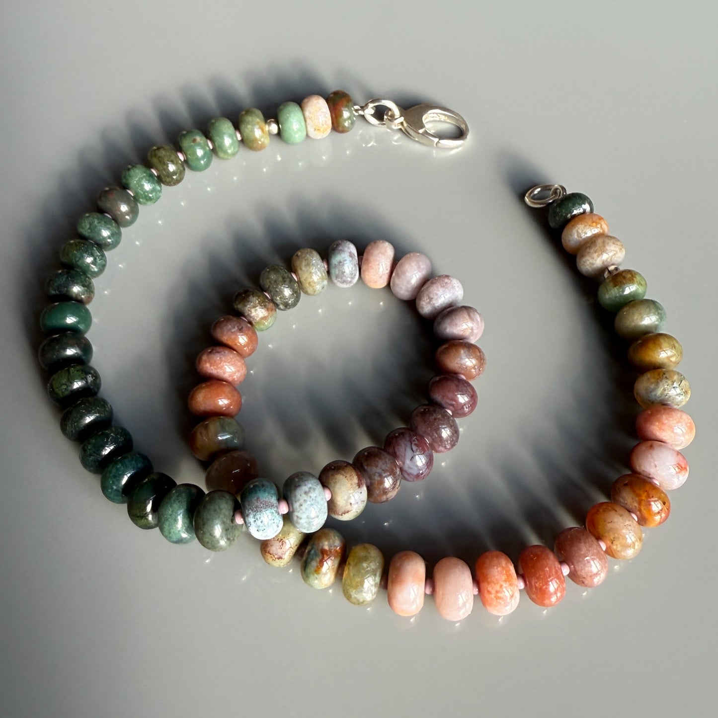 A colorful gemstone necklace with smooth, rounded beads in earthy shades of green, brown, and pink, elegantly arranged and secured with a silver clasp.