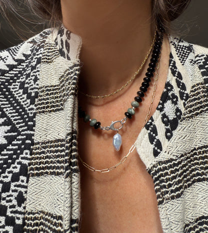A necklace with black beads and a few gray or greenish beads near a silver clasp, featuring a dangling blue crystal or gemstone in the center.