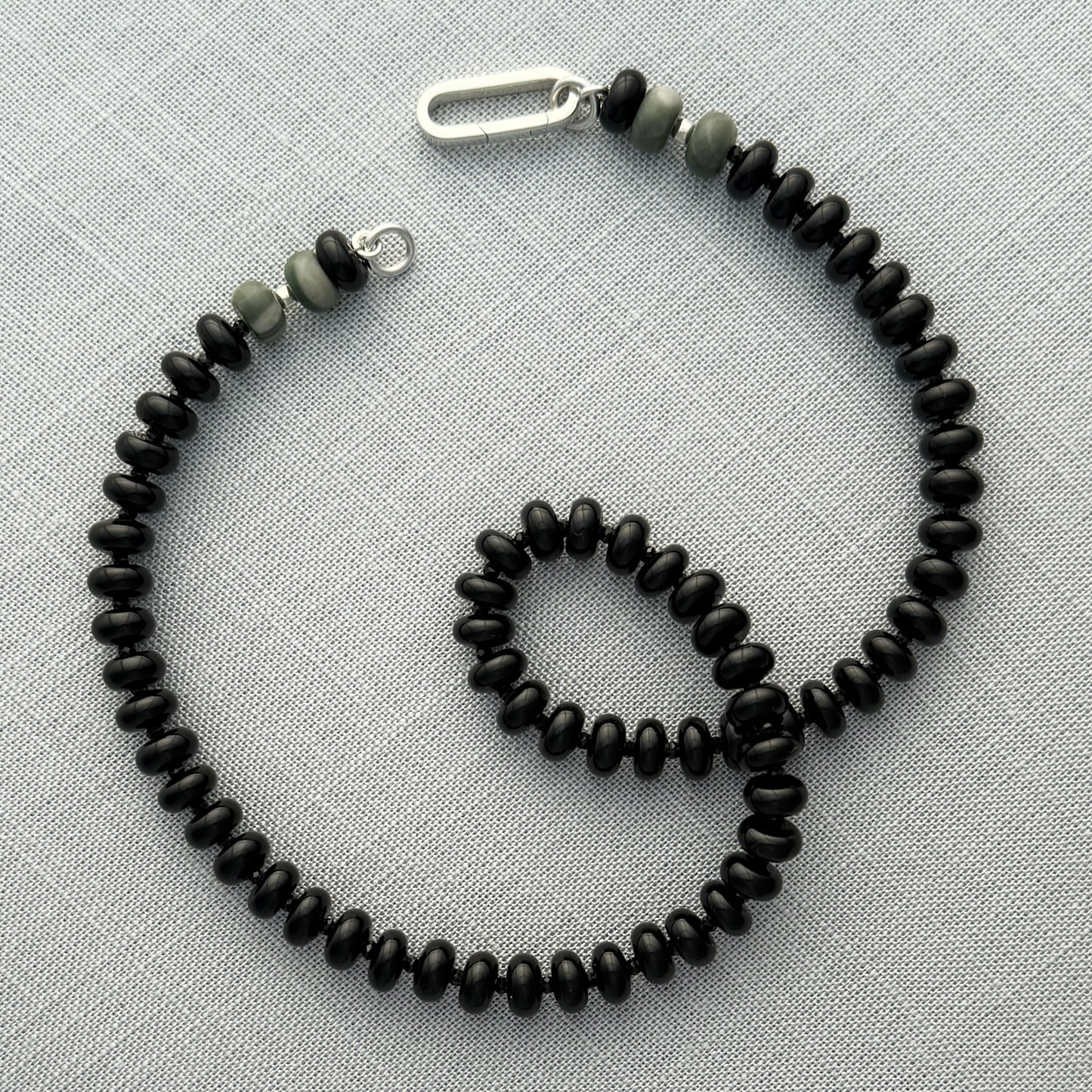 A beaded necklace with black beads and a few grayish-green beads near a silver clasp, laid flat on a textured gray surface.